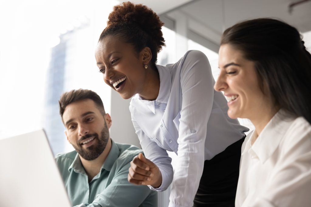 Three colleagues laughing and smiling together as they look at a laptop screen in a bright, modern office.